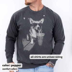 May include: Pepper-colored unisex sweatshirt featuring a graphic of a corgi wearing sunglasses and drinking an iced coffee. The text "all shirts are unisex-sizing" is displayed. The sweatshirt is from Comfort Colors.