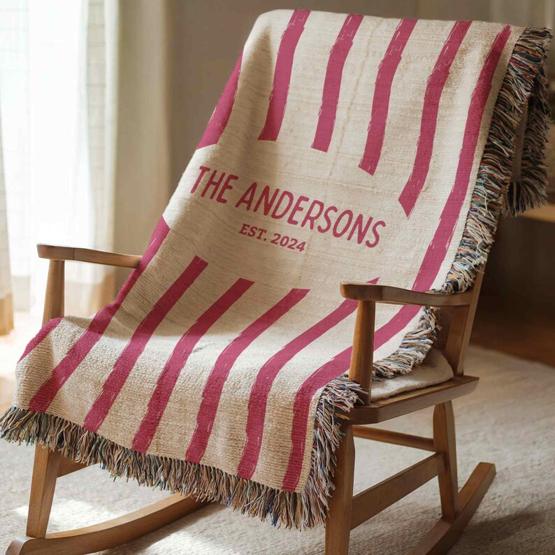 May include: A woven throw blanket with a cream background and bold, vertical magenta stripes. The blanket is draped over a wooden rocking chair and features the text "THE ANDERSONS EST. 2024" in magenta.