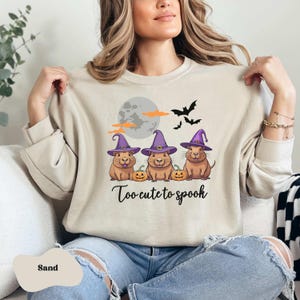 Op de afbeelding: Een beige sweatshirt met een afbeelding van drie cartoon capybara's die toverhoeden dragen en pompoenen vasthouden. De tekst "Too-cute-to-spook" is onder de capybara's gedrukt.
