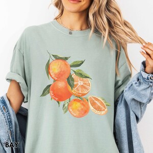 以下が含まれることがあります： 明るい緑色のTシャツに、水彩画で描かれたオレンジが枝についたイラストがプリントされています。オレンジは鮮やかなオレンジ色で、少し黄色がかった色合いをしています。オレンジには「Fruit Trumps」という文字が書かれています。