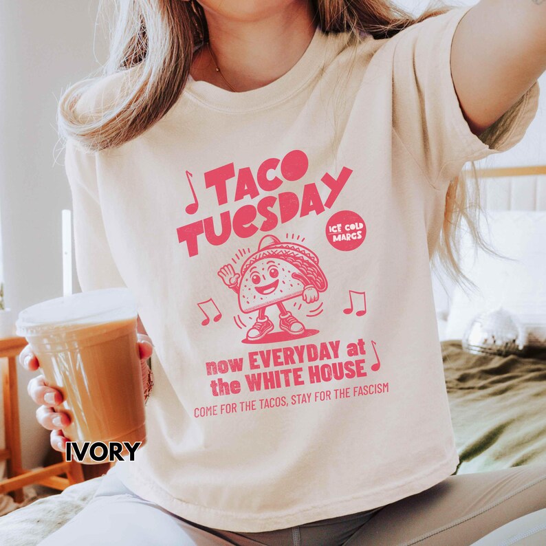 Pode incluir: Camiseta cor marfim com um gr&aacute;fico rosa que diz "TACO TUESDAY" com um personagem de taco de desenho animado. A camisa tamb&eacute;m tem o texto "now EVERYDAY at the WHITE HOUSE" e "COME FOR THE TACOS, STAY FOR THE FASCISM."