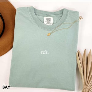 May include: A light green t-shirt with the text "fdt." embroidered on the front. The shirt is made of a soft, comfortable fabric and has a relaxed fit.