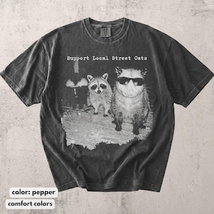 May include: Pepper-colored t-shirt with the text "Support Local Street Cats." The graphic features a raccoon and an opossum wearing sunglasses. The shirt is made of a soft, comfortable material.