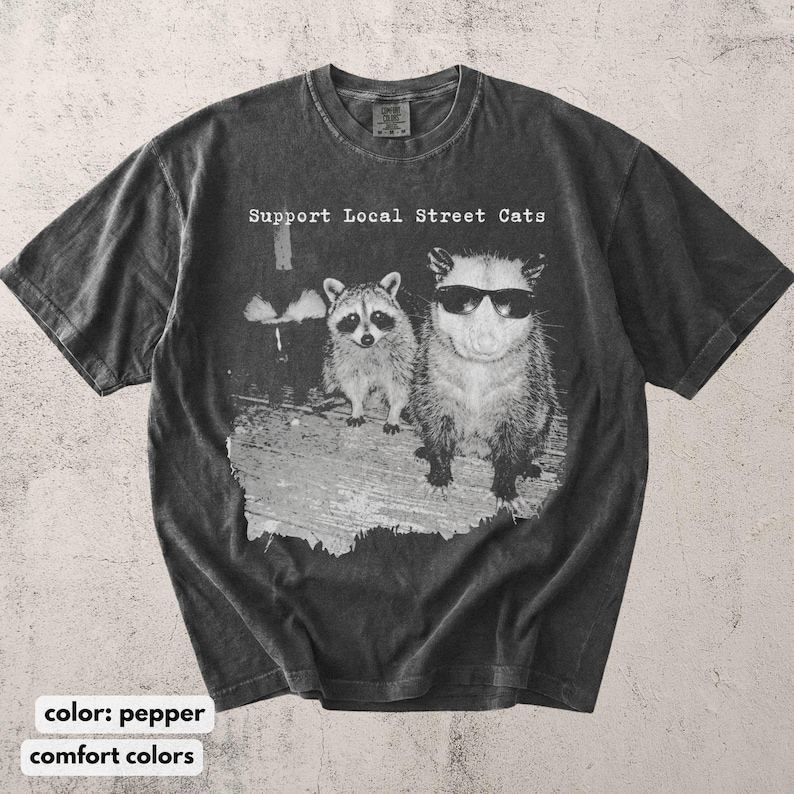 May include: Pepper-colored t-shirt with the text "Support Local Street Cats." The graphic features a raccoon and an opossum wearing sunglasses. The shirt is made of a soft, comfortable material.