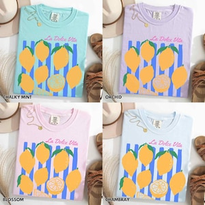 May include: Four pastel-colored t-shirts with the text "La Dolce Vita" and a lemon graphic. The shirts are in chalky mint, orchid, blossom, and chambray colors. Each shirt has a different background color and a similar design of lemons and blue stripes.