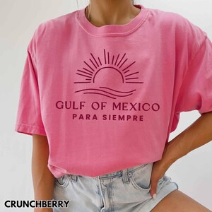 May include: Pink t-shirt with a maroon graphic of a sun setting over waves and the text "GULF OF MEXICO PARA SIEMPRE".