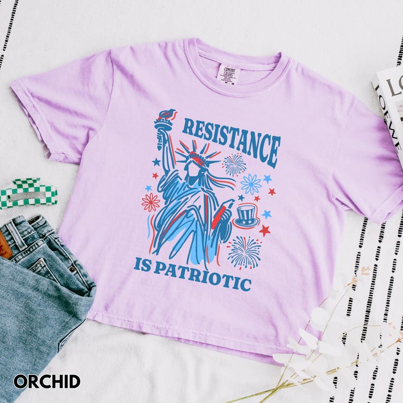May include: Orchid-colored t-shirt with a graphic of the Statue of Liberty holding a torch, with the words "RESISTANCE IS PATRIOTIC" in blue. The shirt has short sleeves and a crew neck. A pair of blue jeans and a green and white hair clip are also visible.