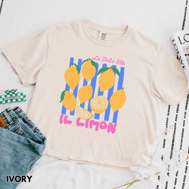 May include: Ivory-colored t-shirt featuring a lemon graphic with the text "La Dolce Vita" and "Il Limon". The design includes yellow lemons, blue stripes, and pink text. The shirt is paired with blue jeans.