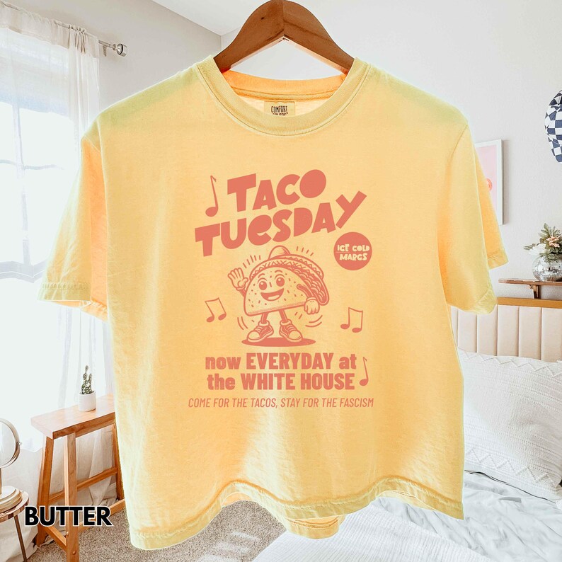 Pode incluir: Camiseta amarelo-manteiga com a inscri&ccedil;&atilde;o vermelha "TACO TUESDAY". O design inclui um personagem de taco de desenho animado e o texto "now EVERYDAY at the WHITE HOUSE". A camiseta est&aacute; pendurada em um cabide de madeira.