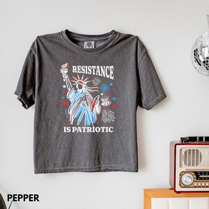 May include: Dark gray t-shirt featuring a graphic of the Statue of Liberty with the text "RESISTANCE IS PATRIOTIC" in red, white, and blue. The design includes fireworks and a top hat. The shirt is short-sleeved.