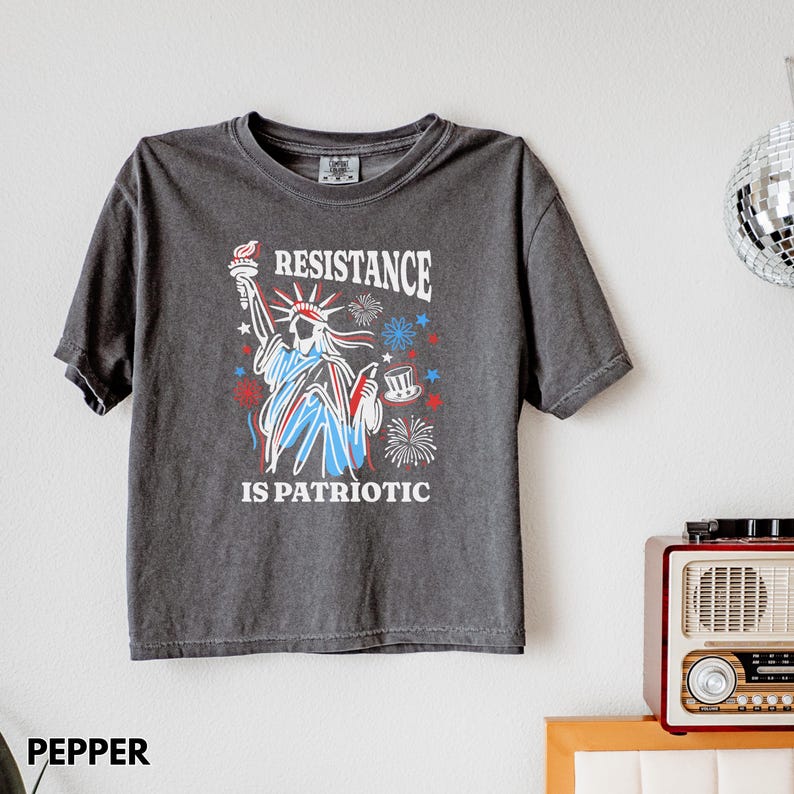 May include: Dark gray t-shirt featuring a graphic of the Statue of Liberty with the text "RESISTANCE IS PATRIOTIC" in red, white, and blue. The design includes fireworks and a top hat. The shirt is short-sleeved.