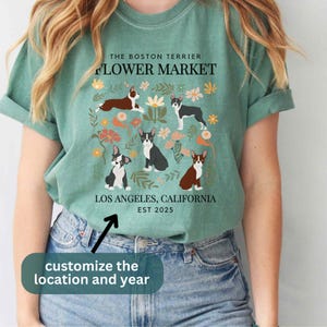May include: A teal blue t-shirt with a floral design featuring Boston Terriers. The text "The Boston Terrier Flower Market" is at the top of the design. The text "Los Angeles, California EST 2025" is at the bottom of the design.