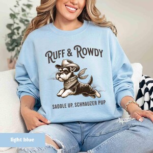 May include: Light blue sweatshirt with a graphic of a miniature schnauzer wearing a cowboy hat and bandana. The text "Ruff & Rowdy" and "Saddle Up, Schnauzer Pup" is printed on the sweatshirt.