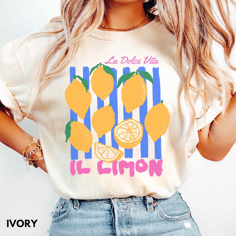 May include: Ivory-colored t-shirt featuring a lemon graphic with the text "La Dolce Vita" and "Il Limon". The design includes yellow lemons, green leaves, and blue vertical stripes. The shirt is a casual wear item.
