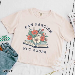 Op de afbeelding: Ivoor kleurig T-shirt met de tekst "BAN FASCISM NOT BOOKS" boven een bloemenillustratie. Het ontwerp toont een open boek met rode, witte en turquoise bloemen. Het shirt wordt gecombineerd met een blauwe jeans.