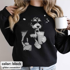 K&ouml;nnte beinhalten: Schwarzes Sweatshirt mit einem Graustufen-Motiv eines Hundes, der eine Sonnenbrille tr&auml;gt und aus einem Becher mit Strohhalm trinkt. Der Text "color: black" und "gildan sweatshirt" stehen unten. Der Hund ist das zentrale Designelement.