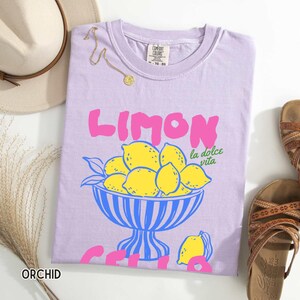 May include: A light purple t-shirt with the word "LIMON" in pink, and a bowl of yellow lemons. The phrase "la dolce vita" is in green. The word "ORCHID" is at the bottom left. A gold necklace and a hat are also in the image.