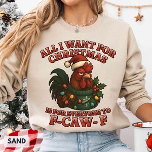 May include: Sand-colored sweatshirt with the text "ALL I WANT FOR CHRISTMAS IS FOR EVERYONE TO F-CAW-F." The graphic features a rooster wearing a Santa hat and a green sweater with Christmas lights. The sweatshirt is a festive holiday design.