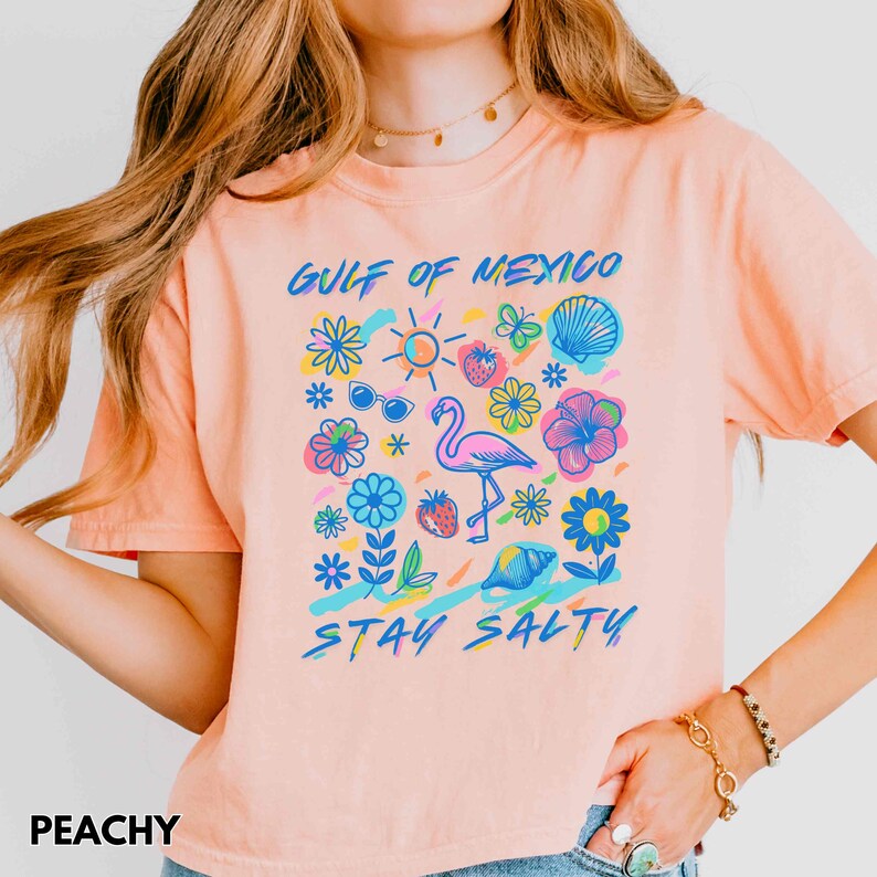 May include: Peach colored t-shirt with a graphic design of a flamingo, flowers, a seashell, sunglasses, and a sun. The text "Gulf of Mexico" and "Stay Salty" are printed on the shirt.
