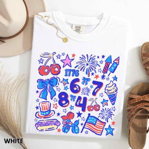 Puede incluir: Camiseta blanca con un dise&ntilde;o gr&aacute;fico colorido que presenta fuegos artificiales, estrellas, cerezas y el n&uacute;mero 1776. El dise&ntilde;o tambi&eacute;n incluye un perrito caliente, un helado y una bandera estadounidense. La palabra "WHITE" est&aacute; impresa en la parte inferior.