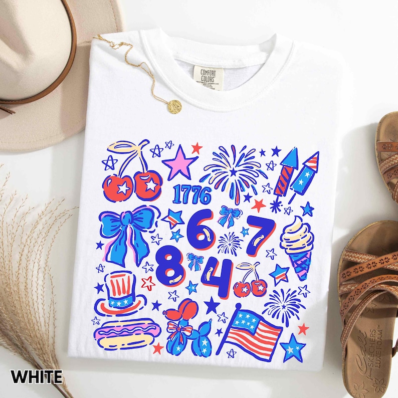 Puede incluir: Camiseta blanca con un dise&ntilde;o gr&aacute;fico colorido que presenta fuegos artificiales, estrellas, cerezas y el n&uacute;mero 1776. El dise&ntilde;o tambi&eacute;n incluye un perrito caliente, un helado y una bandera estadounidense. La palabra "WHITE" est&aacute; impresa en la parte inferior.