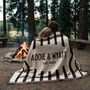 May include: A beige blanket with black vertical stripes and fringed edges. The blanket has the text "ADDIE & WYATT EST. 2022" in black. The blanket is draped over two people sitting near a campfire in a forest.