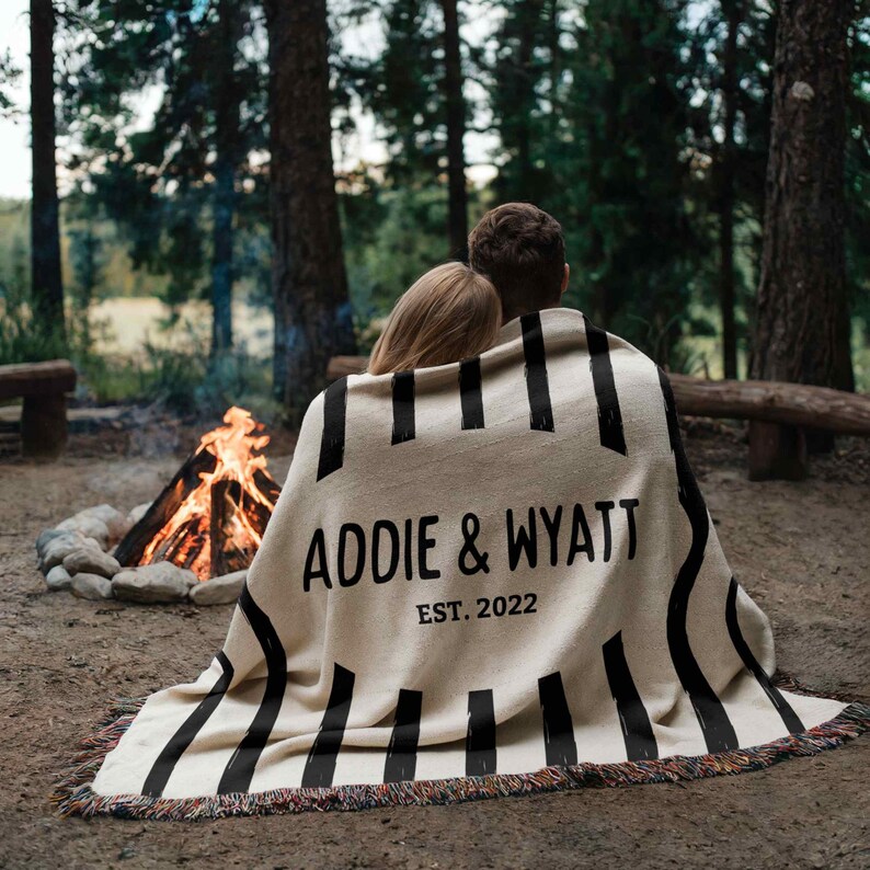 May include: A beige blanket with black vertical stripes and fringed edges. The blanket has the text "ADDIE & WYATT EST. 2022" in black. The blanket is draped over two people sitting near a campfire in a forest.