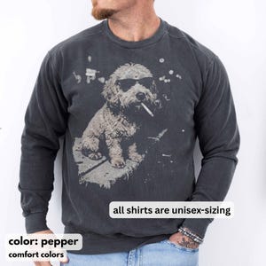 May include: Pepper-colored unisex sweatshirt featuring a graphic of a dog wearing sunglasses and smoking a cigarette. The dog is sitting on a stone surface. The text "all shirts are unisex-sizing" is displayed. The sweatshirt is part of the comfort colors collection.