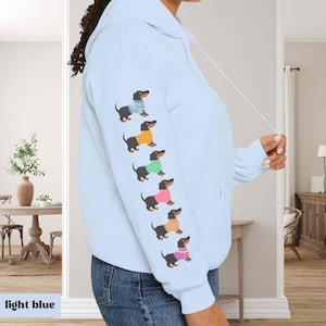 May include: A light blue hoodie with a row of dachshunds wearing colorful sweaters printed on the sleeve. The dachshunds are all facing the right side of the image.