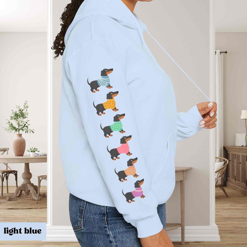 May include: A light blue hoodie with a row of dachshunds wearing colorful sweaters printed on the sleeve. The dachshunds are all facing the right side of the image.