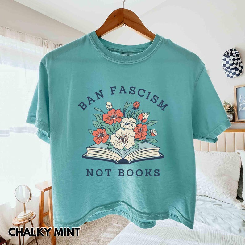 Op de afbeelding: Een mintgroen T-shirt met de tekst "BAN FASCISM NOT BOOKS" boven een open boek en bloemmotief. Het shirt hangt aan een houten hanger. De linkerbenedenhoek toont "CHALKY MINT".