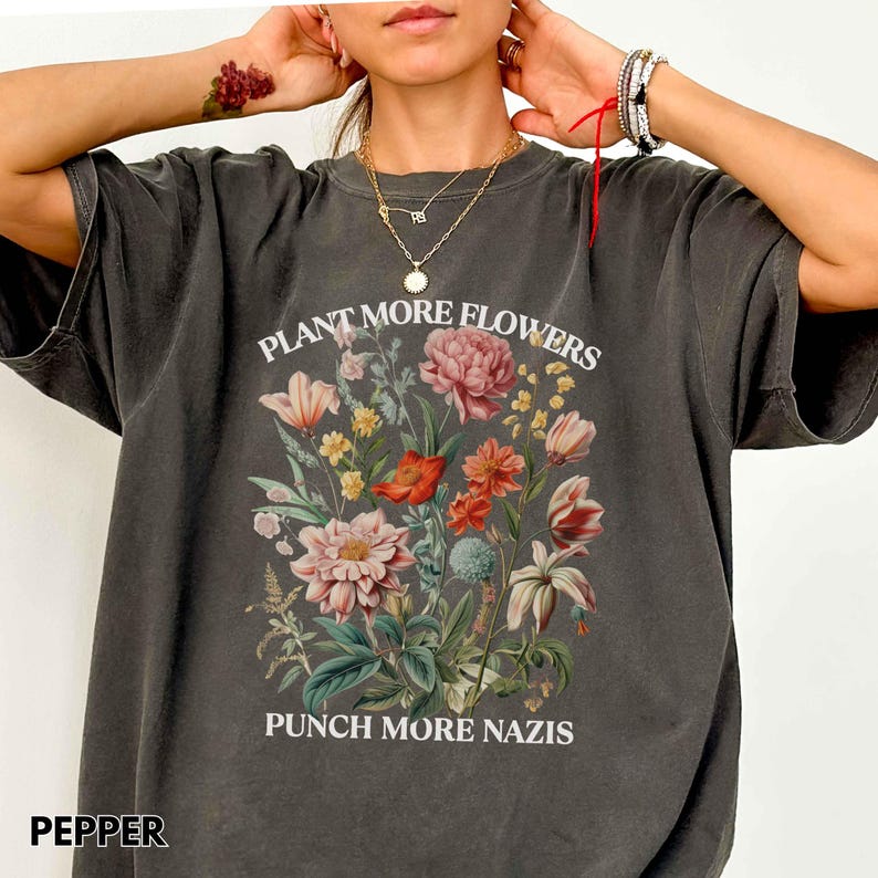 Op de afbeelding: Donkergrijze t-shirt met een bloemenboeket-afbeelding. Het ontwerp bevat roze, rode en gele bloemen, met de tekst "PLANT MORE FLOWERS" erboven en "PUNCH MORE NAZIS" eronder. Het woord "PEPPER" staat linksonder.