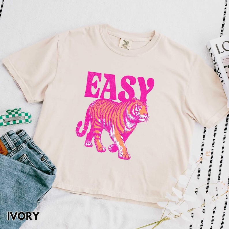 Peut inclure: T-shirt ivoire avec un motif de tigre rose et orange et le mot "EASY" en rose. Le t-shirt est associ&eacute; &agrave; un jean bleu et une pince &agrave; cheveux verte et blanche. Le t-shirt est fait d'une mati&egrave;re douce et confortable.