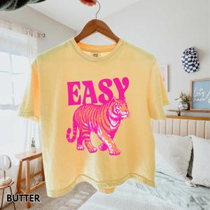 Peut inclure: T-shirt jaune avec le mot "EASY" en lettres roses au-dessus d'un motif de tigre rose et orange. Le t-shirt est suspendu &agrave; un cintre en bois.