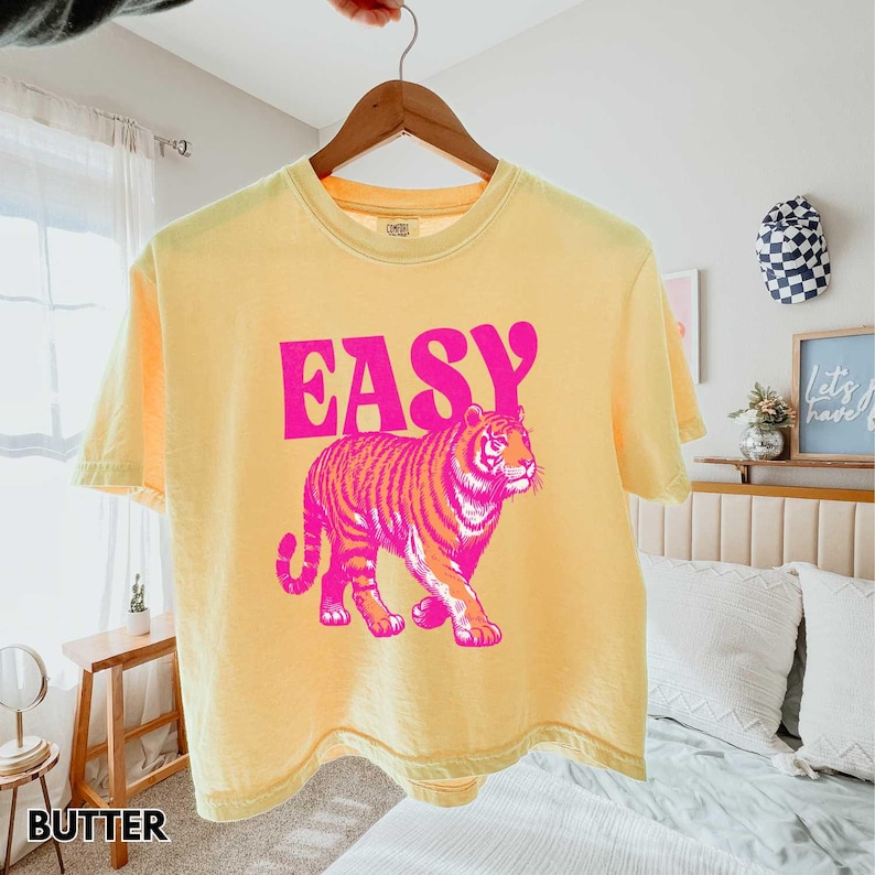 Peut inclure: T-shirt jaune avec le mot "EASY" en lettres roses au-dessus d'un motif de tigre rose et orange. Le t-shirt est suspendu &agrave; un cintre en bois.