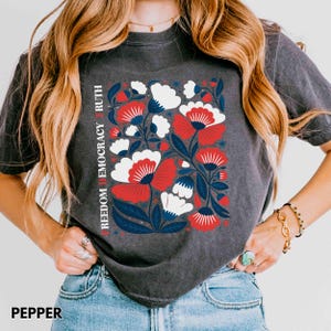 May include: Dark gray t-shirt with a floral design in red, white, and blue. The design includes the words "FREEDOM DEMOCRACY RUTH" vertically on the left side. The word "PEPPER" is at the bottom of the shirt.