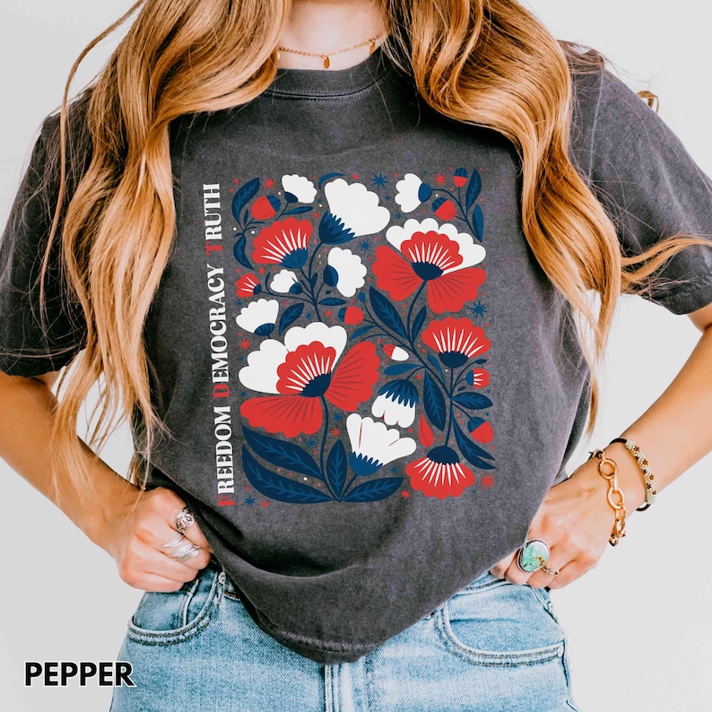 May include: Dark gray t-shirt with a floral design in red, white, and blue. The design includes the words "FREEDOM DEMOCRACY RUTH" vertically on the left side. The word "PEPPER" is at the bottom of the shirt.