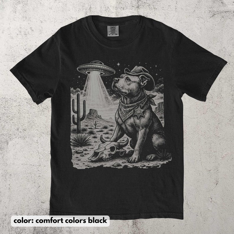以下が含まれることがあります： 黒いTシャツ。カウボーイハットとバンダナをつけた犬がUFOを見上げているグラフィック。デザインには、頭蓋骨とサボテンのある砂漠の風景が含まれています。「color: comfort colors black」の文字が下にあります。