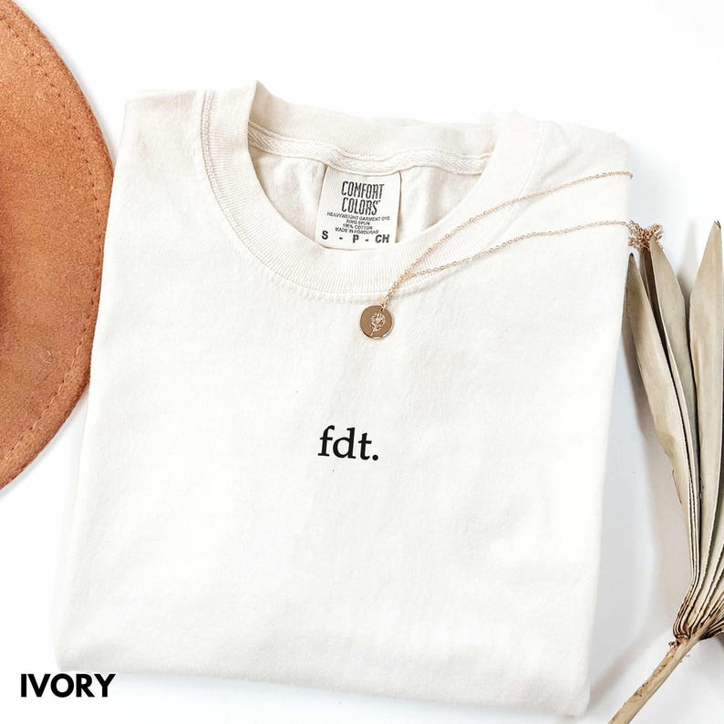 May include: Ivory t-shirt with the text "fdt." embroidered on the front. The shirt is laying flat on a white surface with a brown hat and a dried plant in the background.