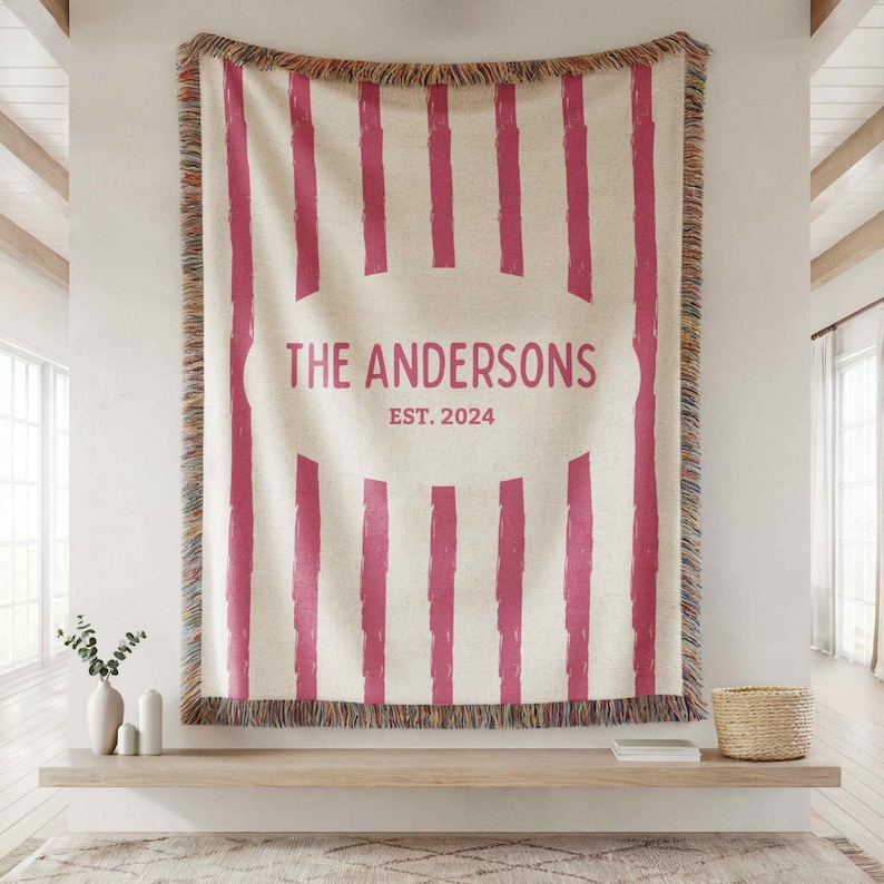 May include: Cream-colored woven throw blanket with vertical magenta stripes and fringed edges. The center features the text "THE ANDERSONS EST. 2024" in magenta. The blanket is displayed on a wall.