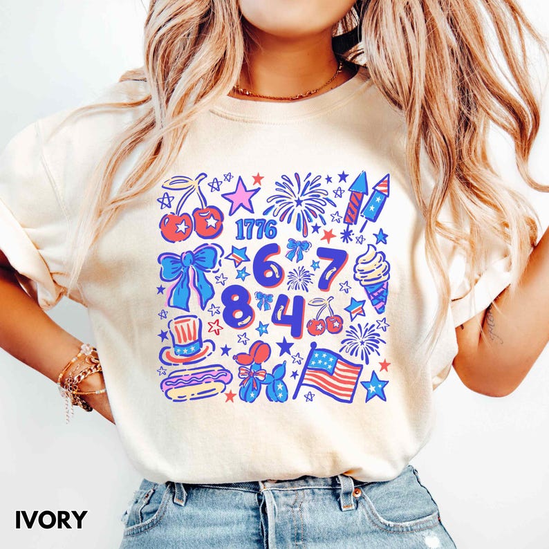 Puede incluir: Camiseta color marfil con cuello redondo y dise&ntilde;o gr&aacute;fico colorido. El dise&ntilde;o incluye el n&uacute;mero 1776, fuegos artificiales, estrellas, cerezas, un lazo, un helado, un perrito caliente, un sombrero de copa, un perro globo y una bandera americana.