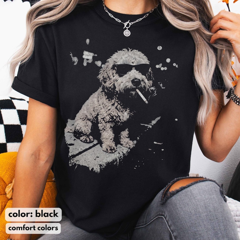 May include: Black t-shirt featuring a grayscale illustration of a dog wearing sunglasses and smoking a cigarette. The dog is sitting on a gray surface. The text "color: black" and "comfort colors" is at the bottom.