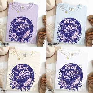 May include: Four t-shirts in different colours, each featuring the same graphic design. The graphic is a circle with a whale, flowers, and the text "Eat the Rich". The t-shirts are in orchid, ivory, white, and Chambray.