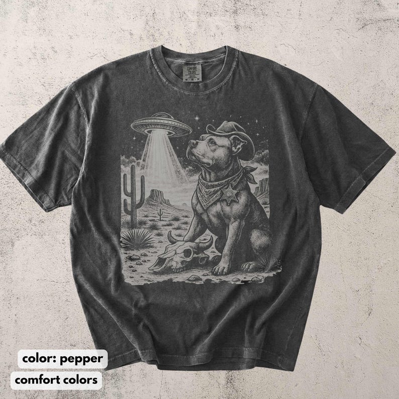 以下が含まれることがあります： コショウ色のTシャツ。カウボーイハットとバンダナを身に着けた犬のグラフィックが特徴で、UFOが砂漠のシーンに光を当てています。犬は頭蓋骨の近くに座っています。「color: pepper」と「comfort colors」の文字が下にあります。