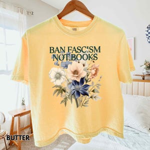 Puede incluir: Camiseta amarilla mantequilla con el texto "BAN FASCISM NOT BOOKS" en verde oscuro. Un ramo floral en tonos blancos, rosas y azules est&aacute; impreso debajo del texto. La camiseta est&aacute; colgada en una percha de madera.