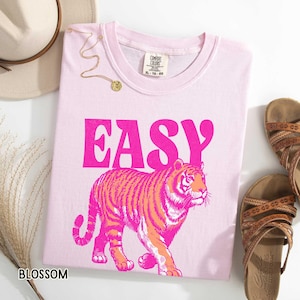K&ouml;nnte beinhalten: Hellrosa T-Shirt mit dem Wort "EASY" in leuchtend pinkfarbenen Buchstaben &uuml;ber einer rosa-orangenen Tigerillustration. Das Shirt ist von der Marke Comfort Colors, Gr&ouml;&szlig;e XL-TG-20.