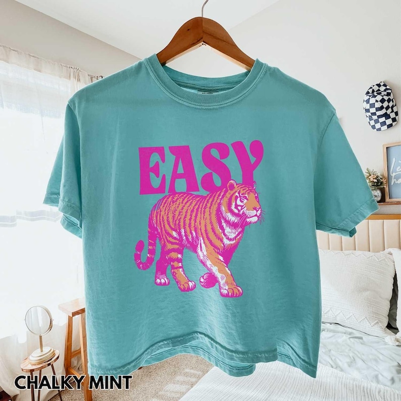 Peut inclure: T-shirt turquoise avec le mot "EASY" en rose, au-dessus d'un motif de tigre rose et orange. Le t-shirt est suspendu &agrave; un cintre en bois.