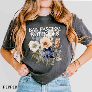 Puede incluir: Camiseta gris oscuro con el texto "BAN FASCISM NOT BOOKS" sobre un ramo floral. El ramo presenta flores blancas, melocot&oacute;n y azules con hojas verdes. La camiseta es de estilo casual y c&oacute;modo.