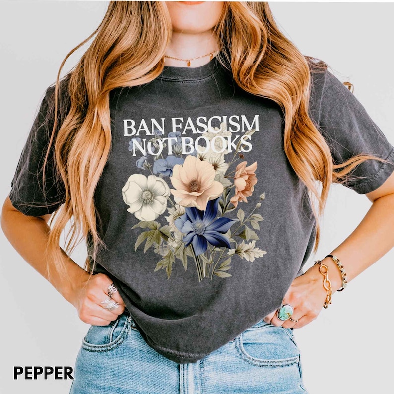 Puede incluir: Camiseta gris oscuro con el texto "BAN FASCISM NOT BOOKS" sobre un ramo floral. El ramo presenta flores blancas, melocot&oacute;n y azules con hojas verdes. La camiseta es de estilo casual y c&oacute;modo.