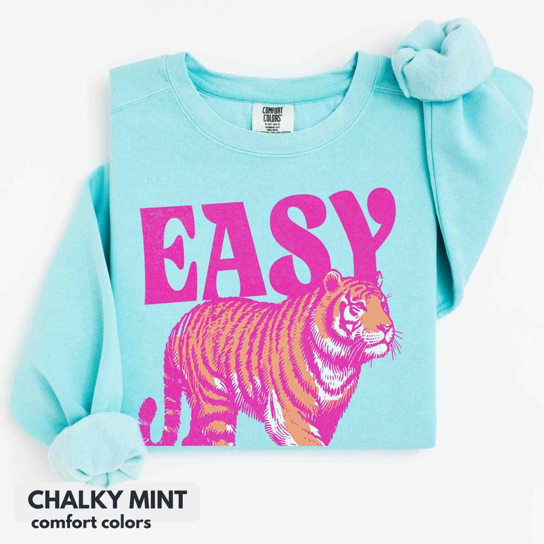 May include: A light blue crewneck sweatshirt featuring the word EASY in large, bold, hot pink letters above a graphic of a tiger rendered in shades of hot pink and orange. The garment has a Comfort Colors brand label at the inner neckline.